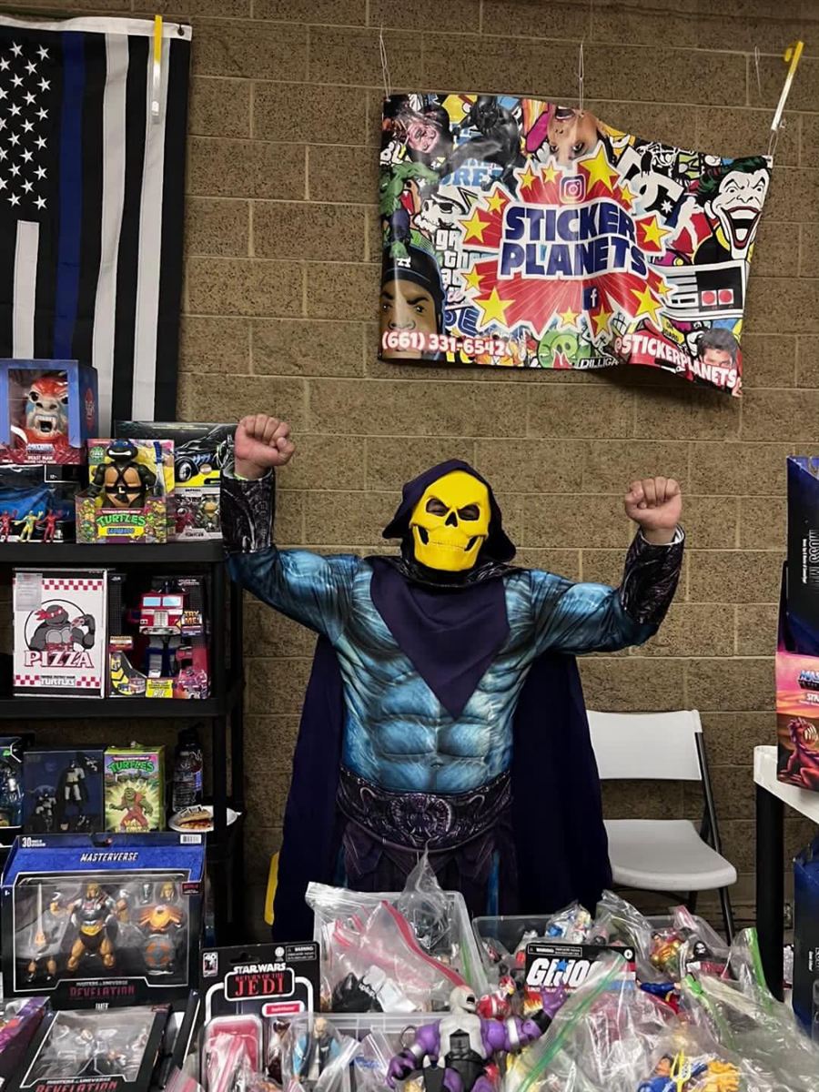 A Skeletor cosplayer celebrating behind a table of toys and collectibles at the October 10 show.